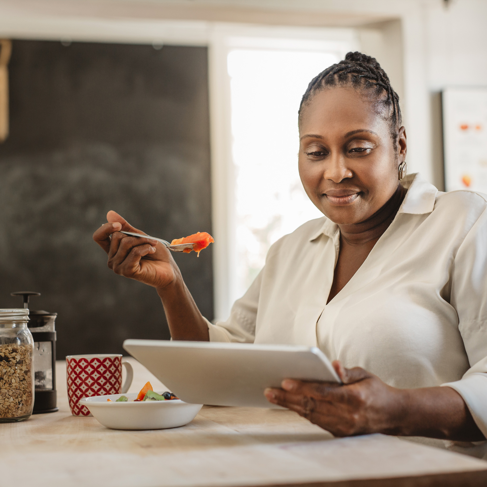 woman sitting in kitchen using a tablet while eating a healthy breakfast