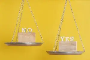 Two balance scales on a yellow background, with the scale on the right labeled ‘YES’ weighing more than the scale on the left labeled ‘NO’.