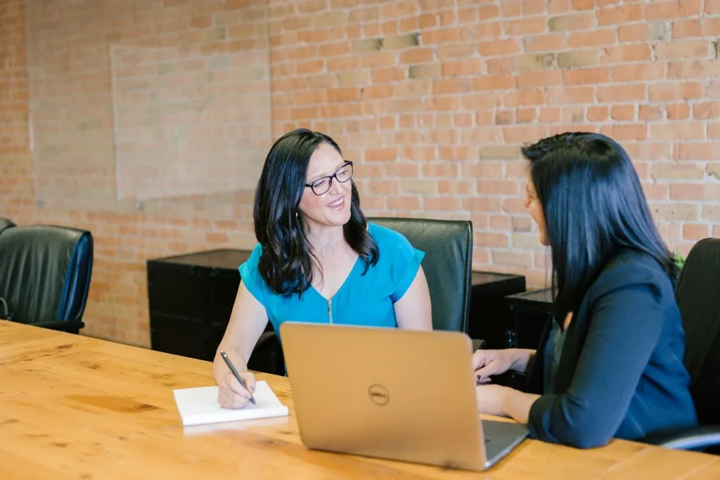 Two women engaged in conversation at a wooden table with a laptop and notepad.
