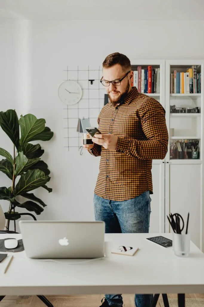 Man in a checkered shirt stands in a modern office, holding a phone and cup, surrounded by shelves.