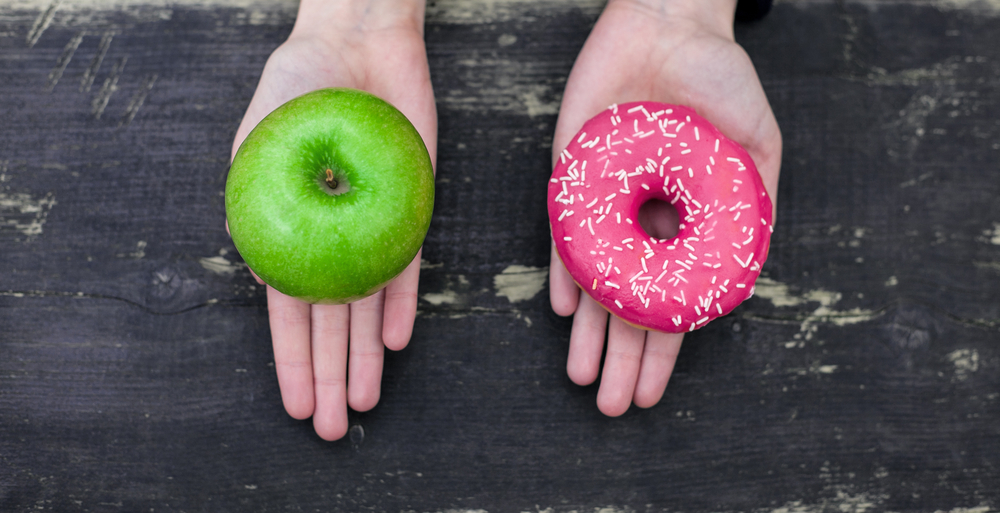 Hands holding a green apple and pink donut, symbolizing choice between healthy and indulgent options