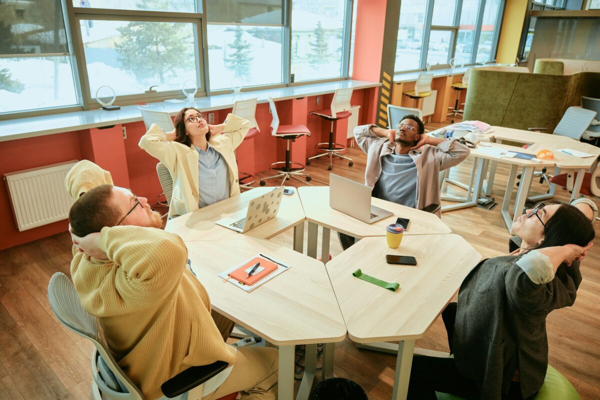 Five individuals relax in an office space, leaning back in chairs with laptops on a table.