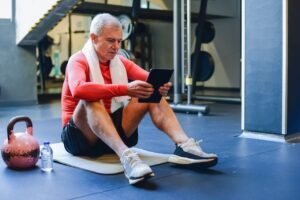 Older adult man using a digital wellness platform on a tablet in a gym, representing health coaching and fitness tools for senior living communities.