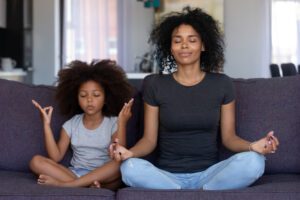 Mother and daughter sitting cross-legged on a couch, practicing meditation together at home, symbolizing movement, mindfulness, and family connection.