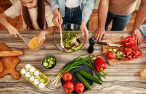 A family prepares a meal together, mixing salad and chopping vegetables on a kitchen counter.
