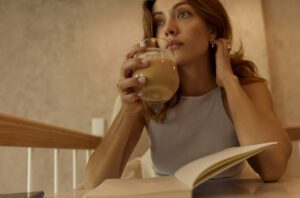 Woman sitting with a drink and open book, reflecting quietly during a normal daily moment, representing internal self-talk and habit mindset.