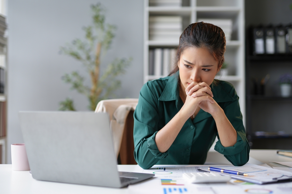 Employee stressed at her desk reviewing financial documents and data on her laptop