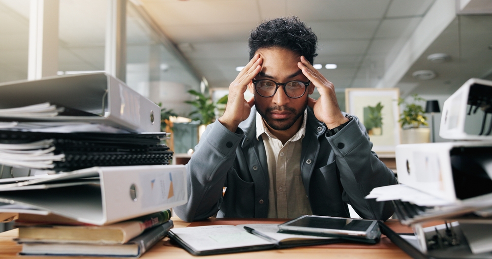 Stressed employee at desk surrounded by binders and paperwork