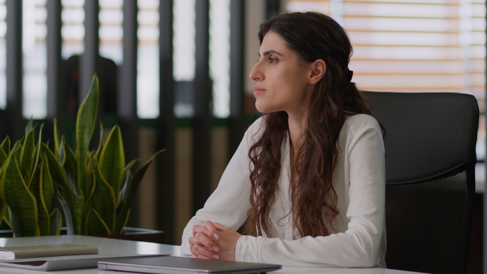 A woman sits at her office desk, hands folded, looking thoughtfully out a window. The image illustrates the quiet presenteeism that often signals caregiver burden in the workplace.