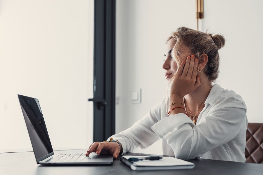 A woman sitting at her desk with her hand on her face, looking distracted and mentally exhausted while working on a laptop