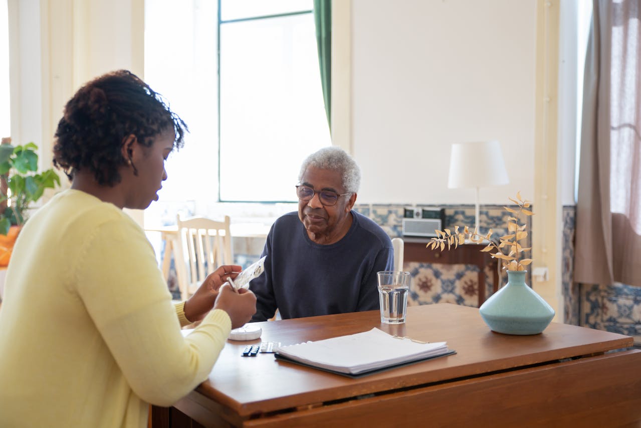 A woman organizes medication at a kitchen table with an older family member, illustrating the home-side of the working caregiver experience.
