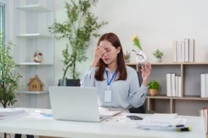Health coach wearing an ID badge rubbing her eyes at a laptop covered in paperwork and reports
