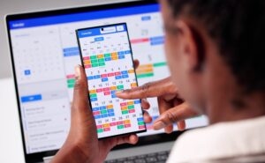 Reviewing a month view calendar on a phone with a scheduling calendar open on a laptop behind it