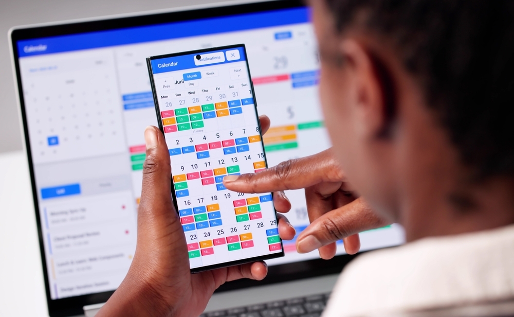Reviewing a month view calendar on a phone with a scheduling calendar open on a laptop behind it