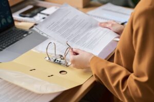 A person filing printed pages into a ring binder at a desk beside a laptop