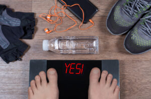 Bare feet on a digital scale reading YES beside sneakers, a water bottle, gloves and a phone