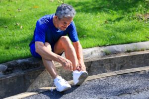 Older man in workout clothes sitting on a curb beside a park lawn, tying his sneakers
