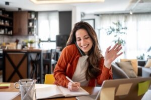 Woman at her kitchen table waving to her health coach on a video call while taking notes