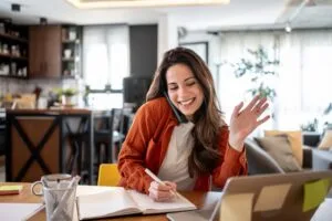 Woman at her kitchen table waving to her health coach on a video call while taking notes