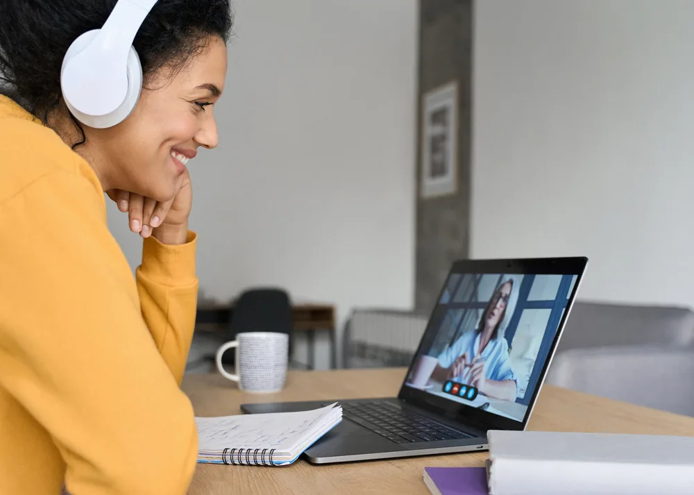 Health coach in headphones smiling during a video call, with a notebook open beside her laptop