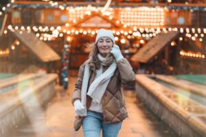 Smiling woman in a winter jacket, knit hat and mittens walking outdoors under string lights at dusk