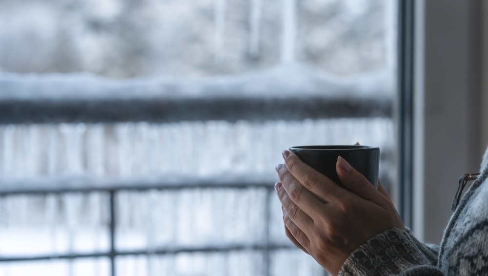 Hands holding a warm mug by a window with snow outside on a gray winter day