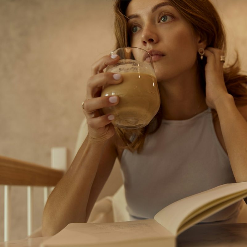 Woman sitting with a drink and open book, reflecting quietly during a normal daily moment, representing internal self-talk and habit mindset.